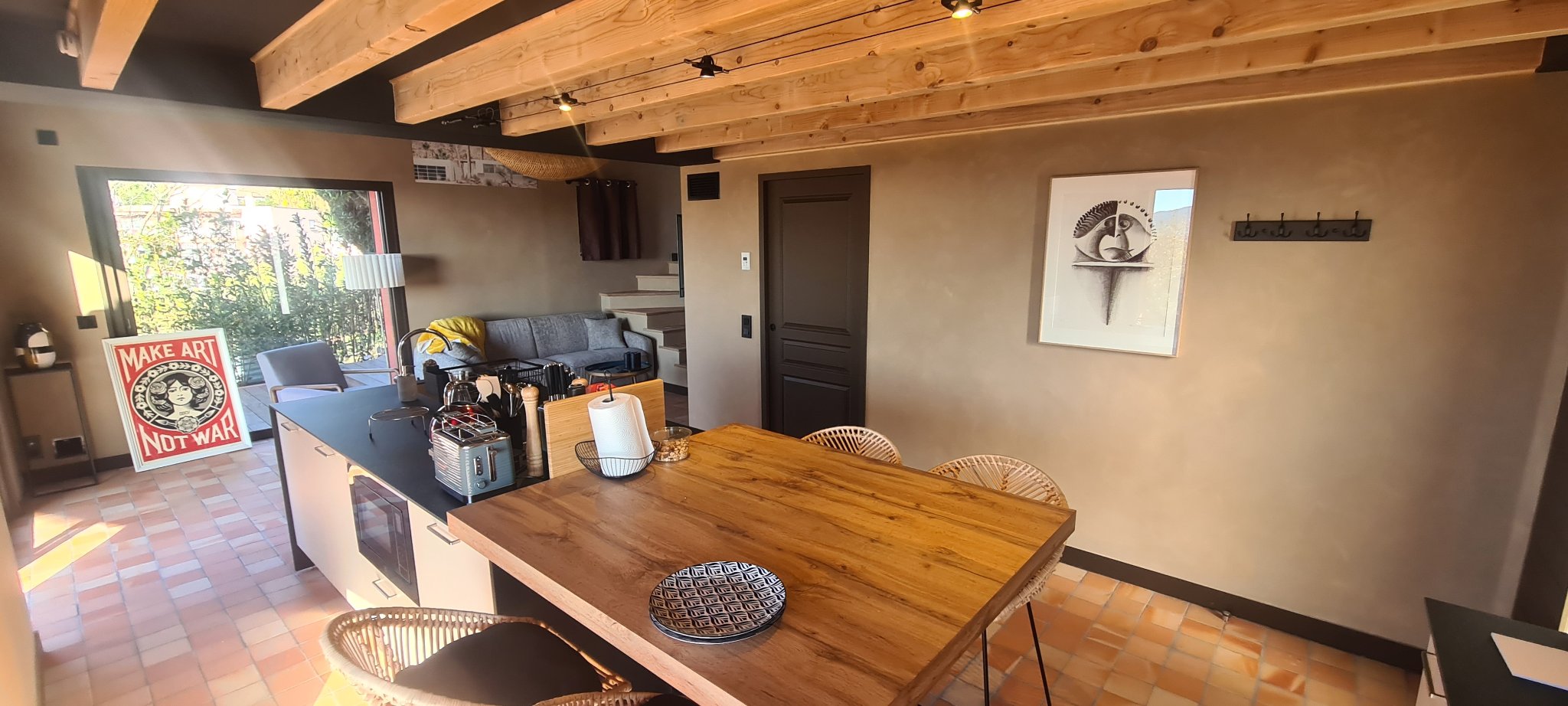 La table à manger en bois avec des chaises en rotin devant la cuisine ouverte, lumière du matin sur la porte-fenêtre de la terrasse