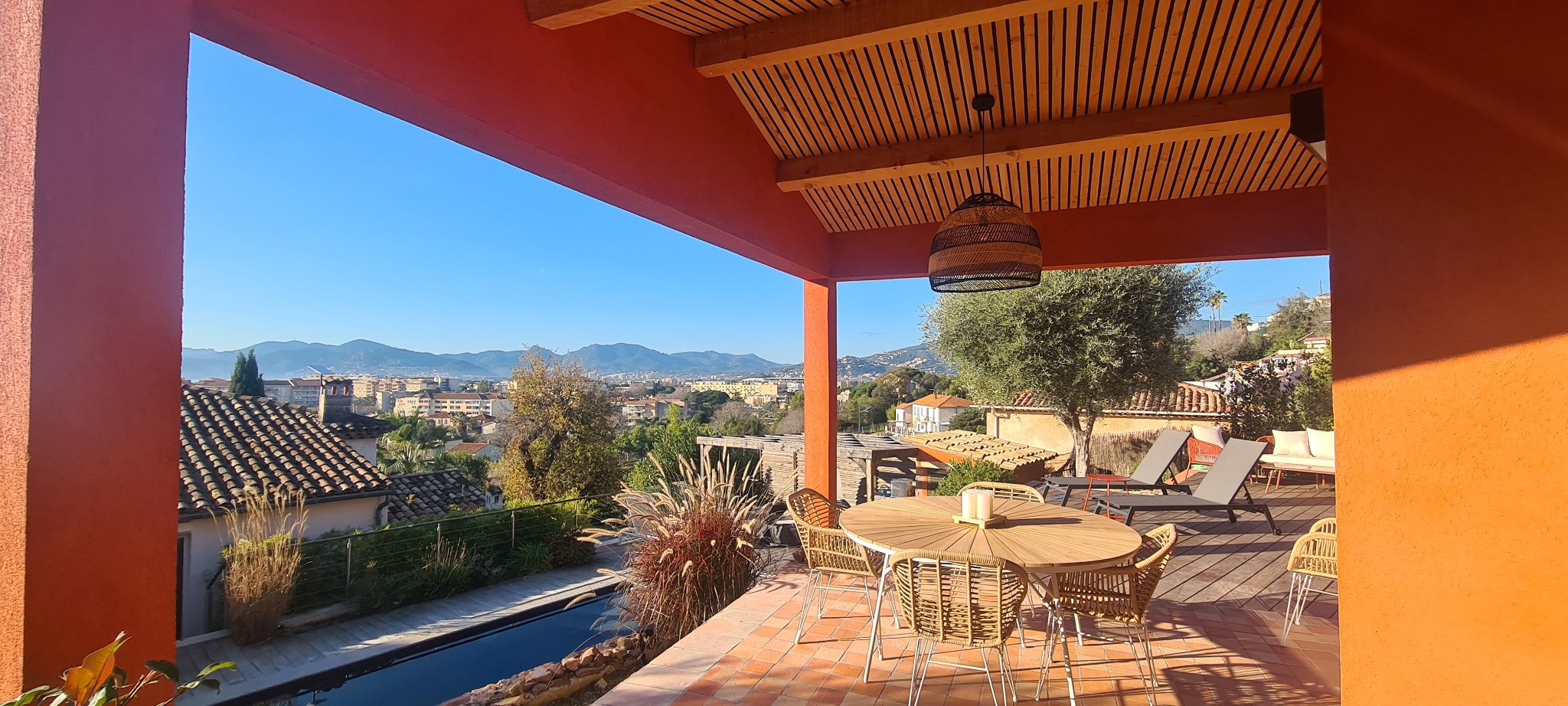The covered terrace of the red house with its round rattan dining table, the pool below and a view over rooftops to Cannes and the Estérel hills