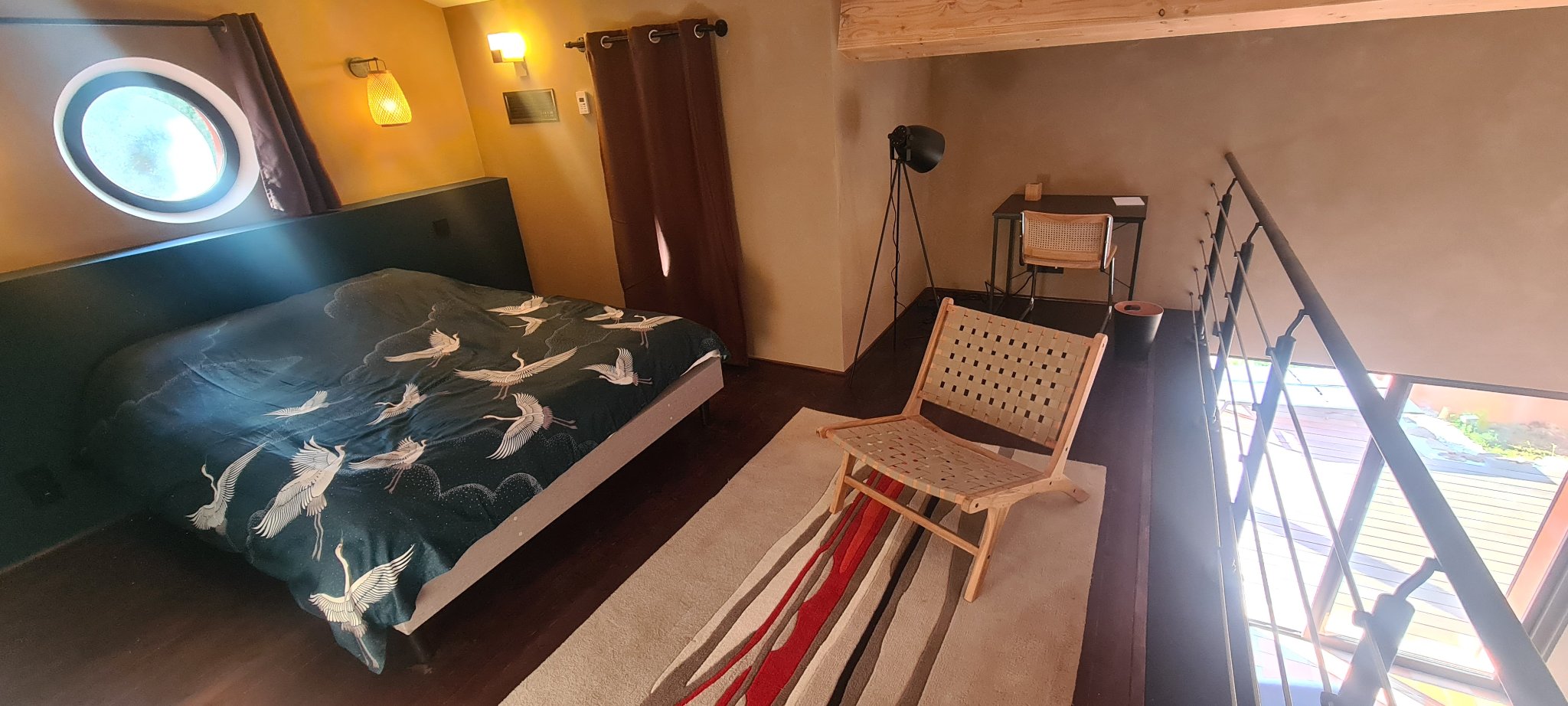 Das Mezzanin-Schlafzimmer mit Doppelbett unter einem runden Fenster, Lesestuhl und Schreibtisch
