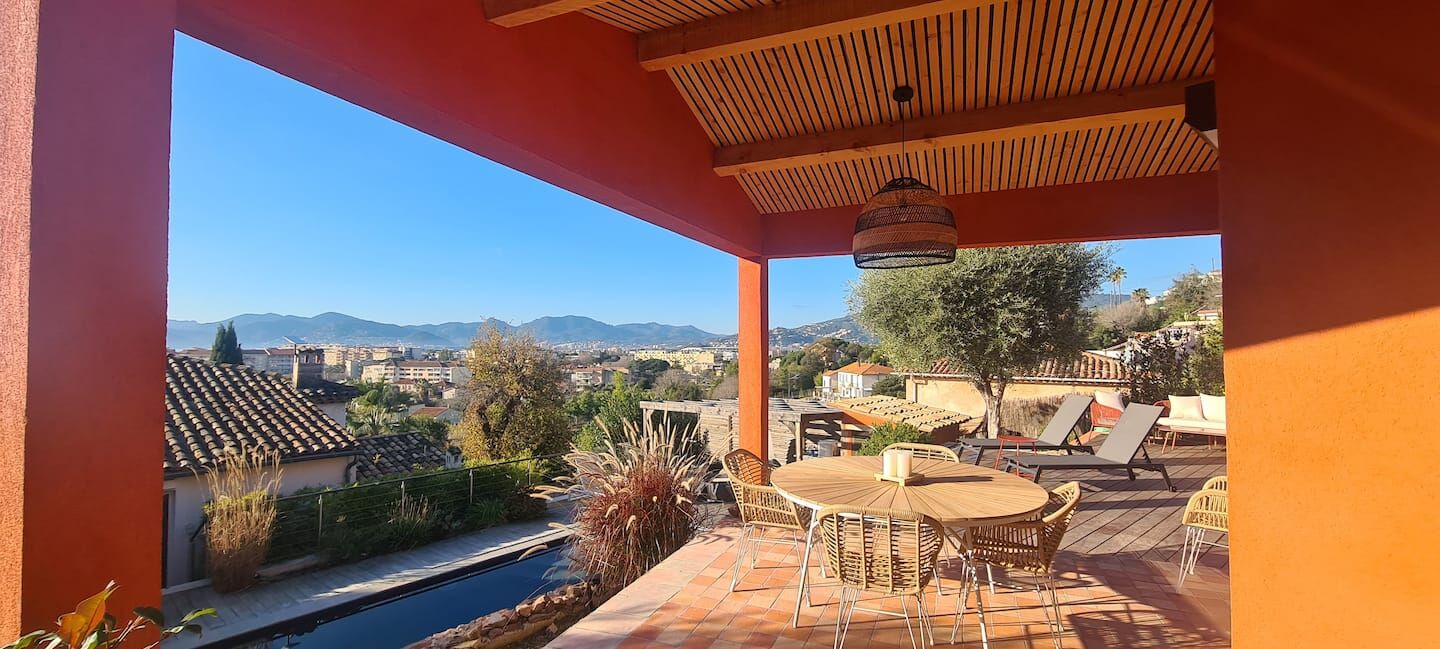 The covered terrace with a round rattan dining table and loungers, the pool and the hills beyond